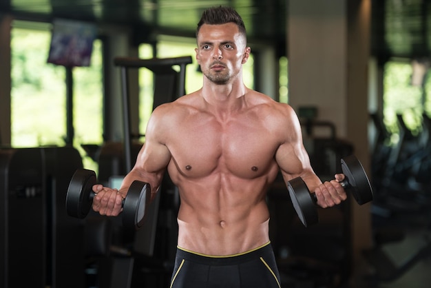 Muskularny młody mężczyzna robi ćwiczenia na biceps z hantlami w nowoczesnej siłowni w centrum fitness