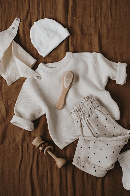 Muślinowy sweter na szelkach biegacze szczotka skarpetki kapelusz akcesoria i zabawki dla noworodków ubrania i zabawki ustawione na brązowym lnianym kocu moda skandynawskie podstawowe artykuły dla noworodka widok z góry na płasko