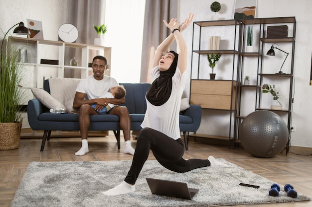 Muzułmańska dama w hidżabie robi trening pilates z laptopem w domu, ćwicząc jogę online