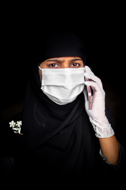 Muzułmańska dziewczyna w masce ochronnej i rękawiczkach rozmawia na swoim smartfonie Kobieta z czarnego hidżabu w niebieskiej masce dla bezpieczeństwa koronawirusa