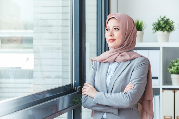 Muzułmańska kierowniczka stojąca przy oknie i radośnie obserwująca widok na zewnątrz budynku. Biznes kobieta relaksująca praca w biurze. Styl życia pani biura.