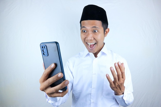 Zdjęcie muzułmański azjat wykazuje podekscytowany wyraz twarzy, trzymając w ręku telefon komórkowy i czytając wiadomość