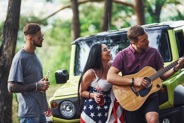 Muzyk gra piosenkę na gitarze. Przyjaciele spędzają miły weekend na świeżym powietrzu przy swoim zielonym samochodzie z flagą USA.