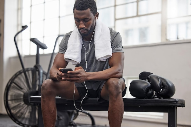 Muzyka i fitness przez telefon z czarnym mężczyzną, który robi sobie przerwę na siłowni podczas treningu bokserskiego Ćwicz w mediach społecznościowych lub aplikacji sportowej z bokserem lub sportowcem śledzącym postępy i odpoczywającym na ławce