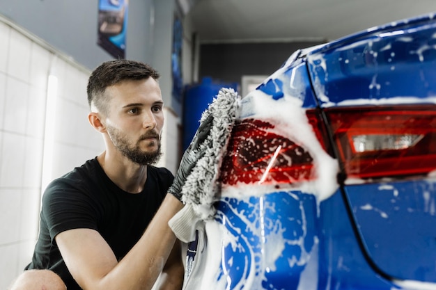 Mycie ręczne w rękawicach z mikrofibry z piankową karoserią w garażu Myjnia samochodowa wykonująca ręczne mycie pianką w serwisie auto detailing