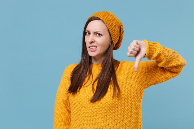 Mylić młoda brunetka kobieta dziewczyna w żółty sweter i kapelusz pozowanie na białym tle na tle niebieskiej ściany portret studio. Koncepcja życia szczere emocje ludzi. Makieta miejsca na kopię. Pokazuje kciuk w dół.