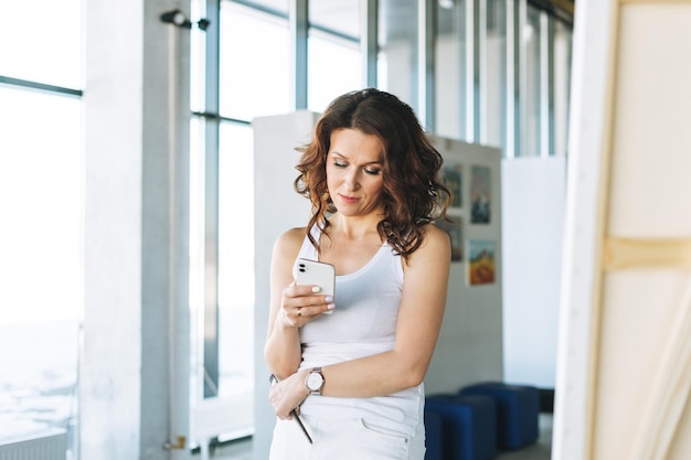 Myśląca artystka w białym ubraniu z pędzlem przy użyciu telefonu komórkowego przed sztalugą w galerii sztuki