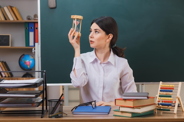 myśląca młoda nauczycielka trzymająca i patrząca na zegarek z piaskiem siedząca przy biurku z szkolnymi narzędziami w klasie