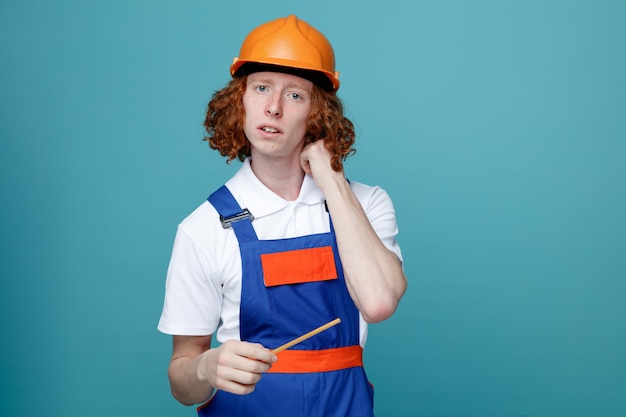 Myślący młody budowniczy mężczyzna w mundurze trzymający ołówek na białym tle na niebieskim tle