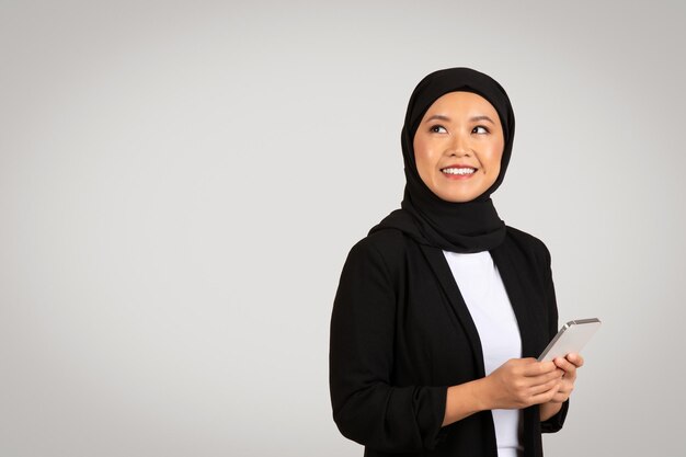 Myślliwa muzułmańska kobieta w czarnym hidżabie i blazerze patrzy w górę trzymając smartfon