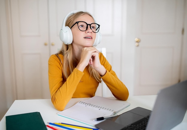 Myślliwa nastolatka w pomarańczowej koszuli słucha uważnie z słuchawkami w biurze domowym