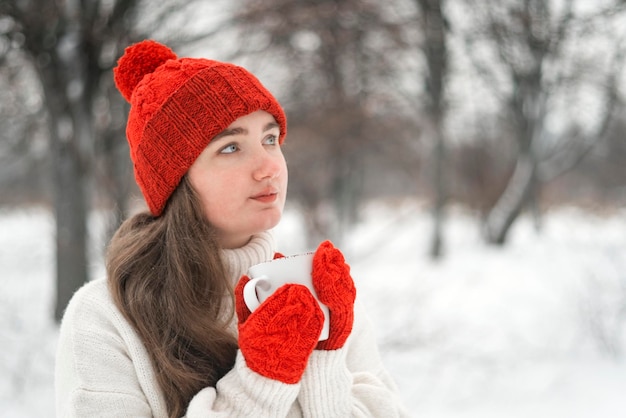 Myślna młoda kobieta w zimowym parku z filiżanką w ręku coś pić Dziewczyna w czerwonym dzianinowym kapeluszu i rękawiczkach na zimę na zewnątrz