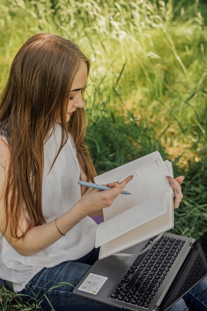 Na banerze młoda dziewczyna pracuje z laptopem na świeżym powietrzu w parku siedząc na trawniku Koncepcja pracy zdalnej Pracuj jako freelancer Dziewczyna bierze udział w kursach na laptopie i uśmiecha się