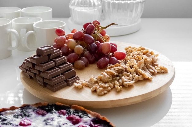 Na białym stole leżą czerwone winogrona, orzechy włoskie, czekoladki i ciasto