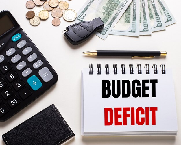 Zdjęcie na białym stoliku kalkulator, kluczyk do auta, gotówka, długopis i notes z napisem deficyt budżetu