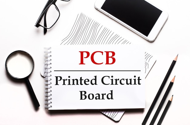 Na białym tle okulary, lupa, ołówki, smartfon i notatnik z napisem PCB Printed Circuit Board