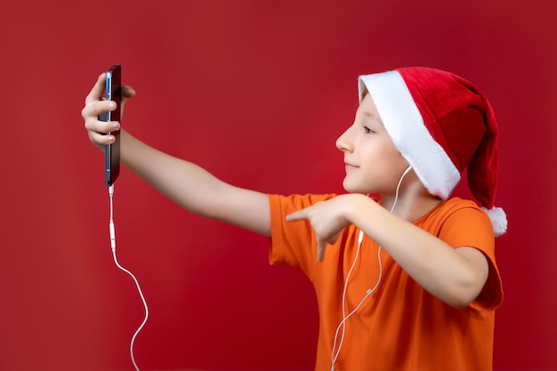 Na czerwonym, świątecznym tle chłopiec w żółtej koszulce trzymający przed sobą telefon i robiący selfie