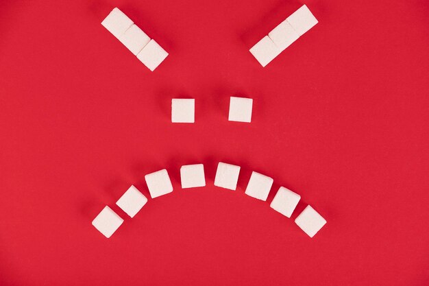 Zdjęcie na czerwonym tle kostki białego cukru w formie złej emotikony. skopiuj miejsce.