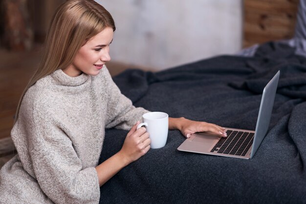 Na produktywny dzień. Młoda kobieta całkiem radosna za pomocą laptopa podczas picia kawy i uśmiechnięty w sypialni.