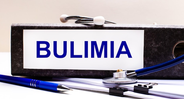 Na pulpicie znajduje się stetoskop, niebieski długopis i szara teczka z tekstem BULIMIA Medical concept