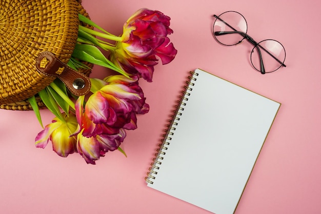 Na różowym tle widnieją okulary do długopisu zeszyt i tulipany w tkanej słomkowej torbie