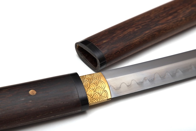 Na rysunku w zbliżeniu przedstawiono japoński miecz z rękojeścią i drewnianą pochwą, zwany Shirasaya, a u nasady ostrza mosiężna pochwa zwana habaki, służy do zaciśnięcia miecza. Nieostrość. Na białym BG.