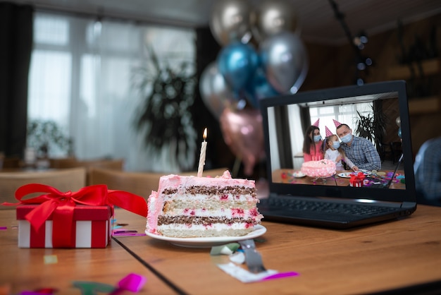 Na stole obok laptopa stoi duży kawałek ciasta i świeca. Rodzina świętuje w Internecie