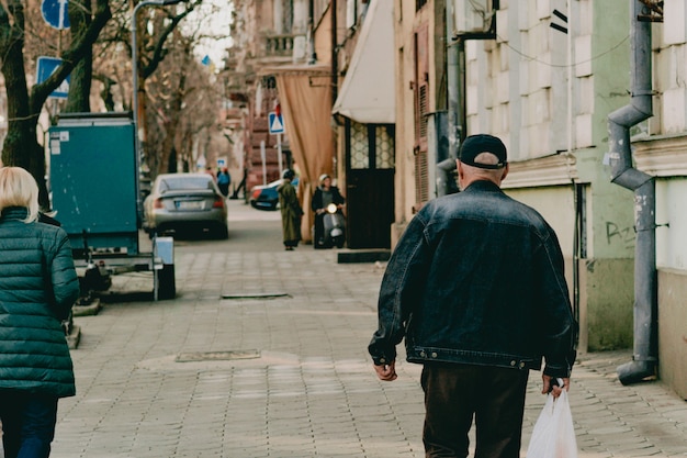 Na ulicę idzie stary człowiek w czapce i dżinsowej kurtce z białą plastikową torbą