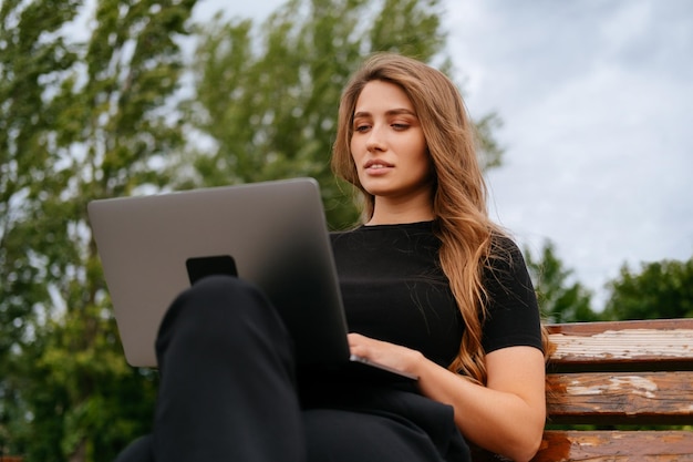 Na zewnątrz portret blondynki młodej kobiety siedzącej na ławce pracującej na laptopie