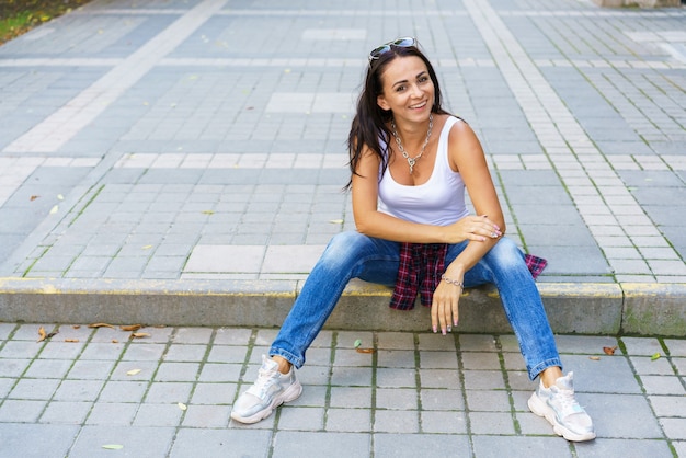 Na zewnątrz portret moda styl życia zadowolony młoda kobieta siedzi na zewnątrz. Ubrana w niebieskie dżinsy i białą koszulkę, uśmiechnięta, szczęśliwa i ciesząca się życiem