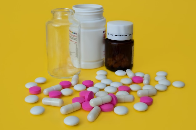 Na żółtym tle białe i różowe tabletki są rozproszone i są tam trzy butelki ze szkła i plastiku. Widok z boku.