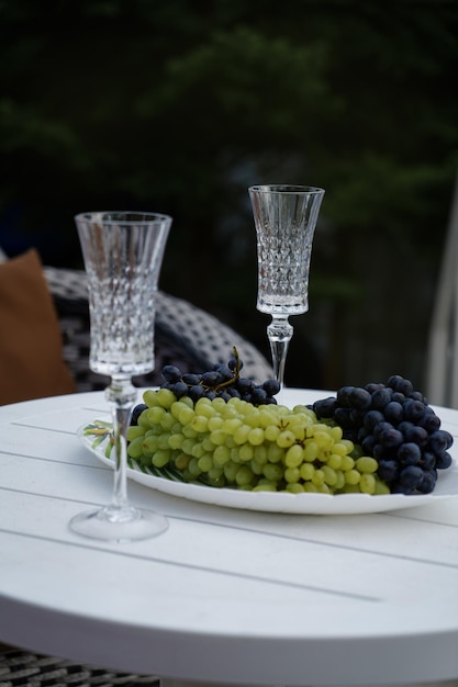 Naczynie z winogronami i kryształową szklanką stoi na białym stole na świeżym powietrzu w letnim domku