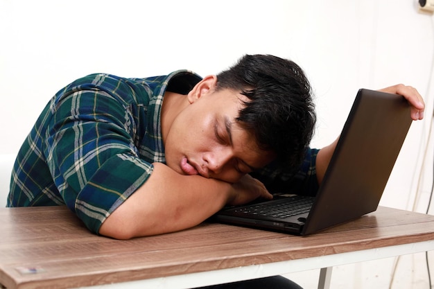 Nadmiernie pracowity młody Azjat śpi, siedząc z laptopem na biurku.
