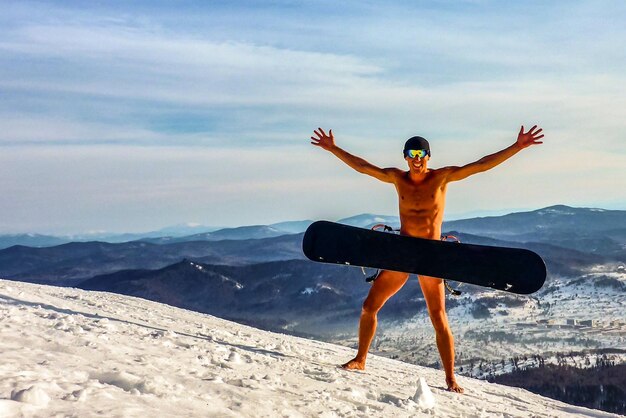 Zdjęcie nagi mężczyzna z deską snowboardową stojący na pokrytej śniegiem górze