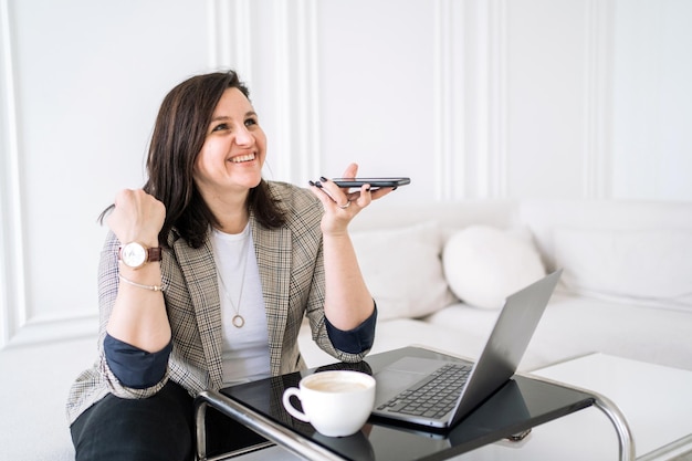 Nagrywa wiadomość głosową, wesoła kobieta korzysta z telefonu i laptopa, freelancer pracuje w domu