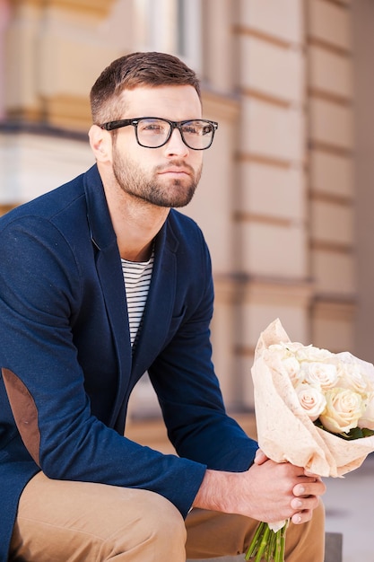 Najświeższe róże dla niej. Przystojny młody mężczyzna w eleganckiej kurtce z bukietem kwiatów i odwracając wzrok siedząc na ławce na świeżym powietrzu