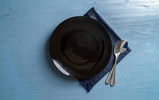 nakrycie stołu tło pusty czarny talerz widelec i łyżka na jasnoniebieskim drewnianym stole z ciemnoniebieskim obrusem widok z góry makieta