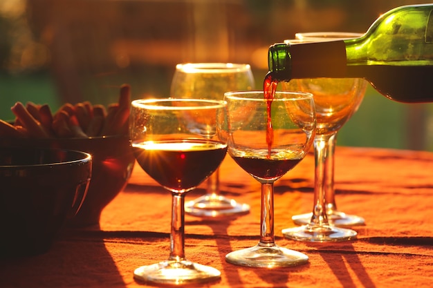 Nalewanie czerwonego wina z butelki do szklanek na stole z tłem przekąsek podczas zachodu słońca