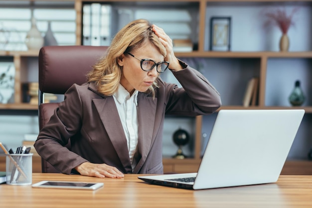 Napięta starsza bizneswoman w okularach wygląda na zmartwioną podczas pracy na laptopie w dobrze wyposażonym pomieszczeniu