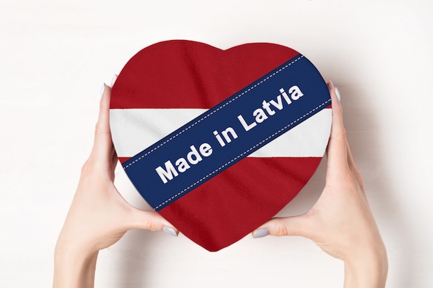 Napis Made in Latvia, flaga Łotwy. Kobiece ręce trzyma pudełko w kształcie serca. .