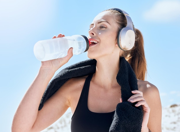 Napój kobiety i butelka wody podczas biegu fitness na świeżym powietrzu z ręcznikiem i muzyką ze słuchawkami lub podcastem strumieniowym dla motywacji do ćwiczeń Nawodnienie wellness i woda pitna po treningu
