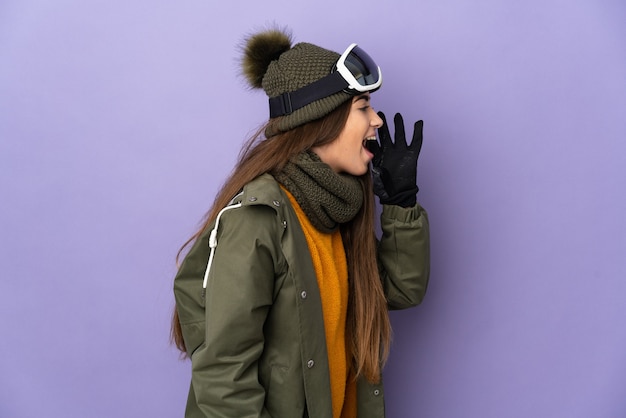 Narciarz kaukaski dziewczyna w okularach snowboardowych na białym tle na fioletowym tle, krzycząc z ustami szeroko otwartymi na bok