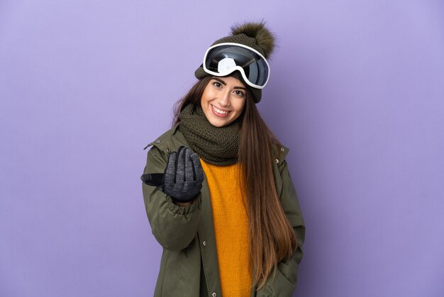 Narciarz kobieta w okularach snowboardowych na białym tle na fioletowej ścianie zapraszając do przyjścia z ręką. Cieszę się, że przyszedłeś