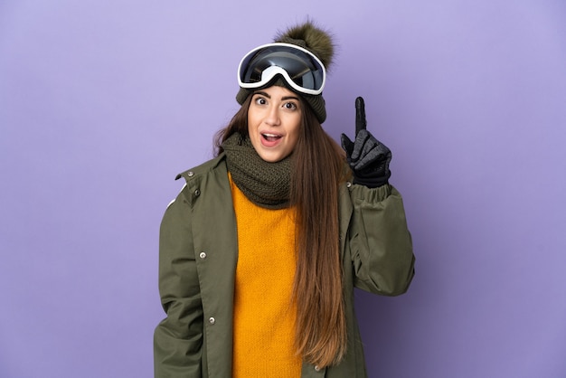 Narciarz kobieta w okularach snowboardowych odizolowanych na fioletowej ścianie, zamierzająca wykonać rozwiązanie, podnosząc palec do góry