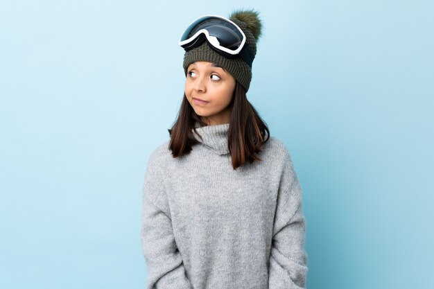 Narciarz rasy mieszanej dziewczyna w okularach snowboardowych na niebieską ścianą, co gest gest patrząc w stronę