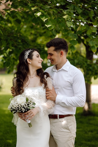 Narzeczeni stoją w parku, trzymając bukiet białych kwiatów.