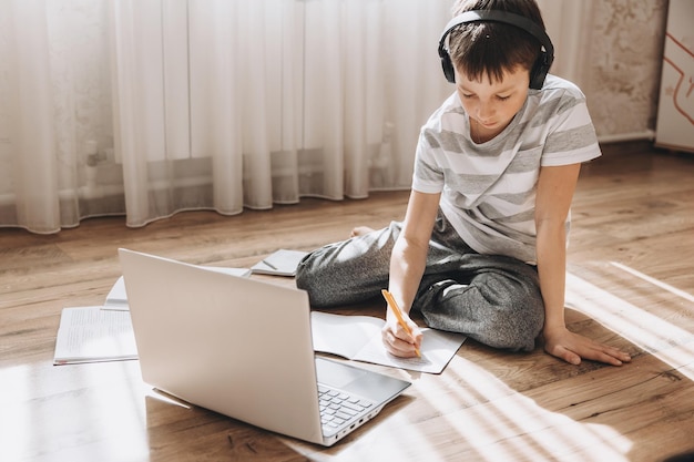 Nastolatek chłopiec w bezprzewodowych słuchawkach leżącego na podłodze z laptopem o wideorozmowy i lekcja online w domu Uczeń studiuje pracę domową Kształcenie na odległość koncepcja szkolenia zdalnego