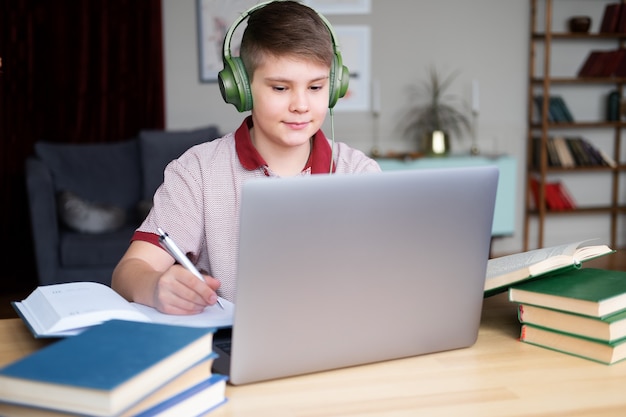 Nastolatek chłopiec w słuchawkach studiuje online za pomocą pisania laptopa w notesie.