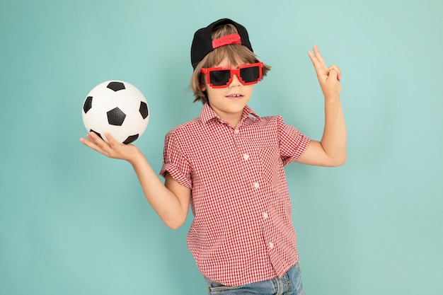 nastolatek chłopiec z czapką, okulary przeciwsłoneczne uśmiecha się i trzyma piłki nożnej na białym
