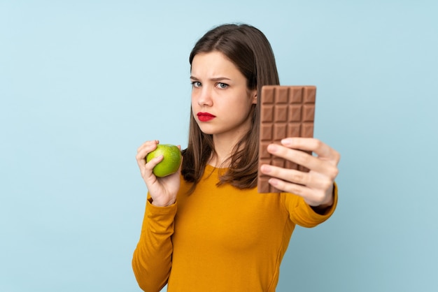 Nastolatek dziewczyna odizolowywająca na błękit ścianie bierze czekoladową pastylkę w jednej ręce i jabłko w drugiej